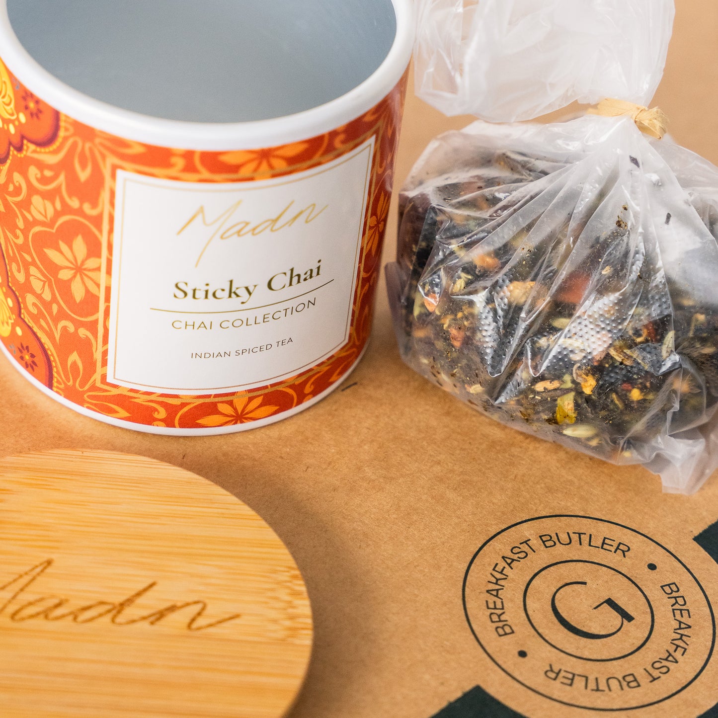 A cylindrical container of 'Madn Sticky Chai' with an ornate orange and red floral pattern, featuring a wooden lid. The label reads 'Madn Sticky Chai, Chai Collection, Indian Spiced Tea,' placed on a wooden surface with a neutral background.