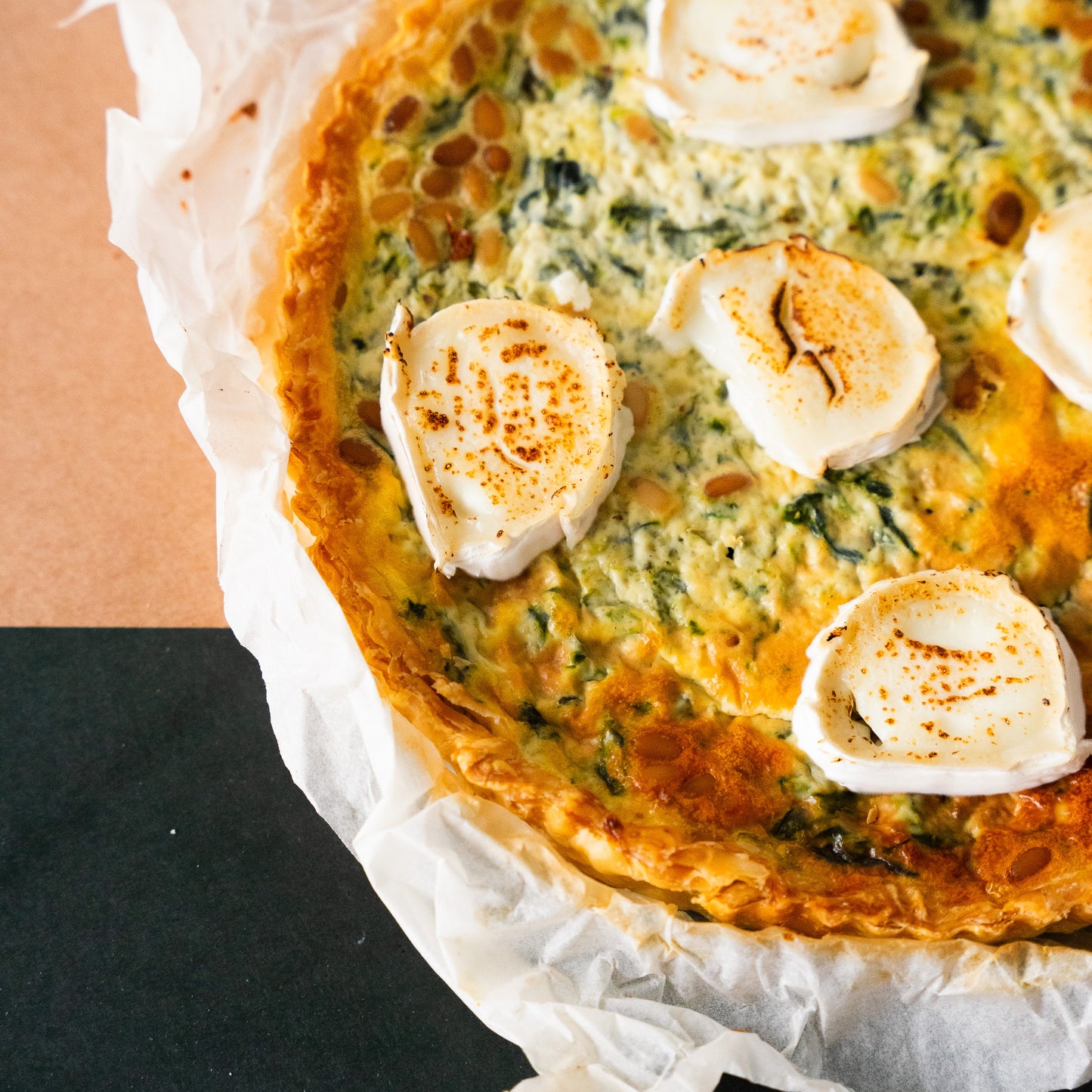 Close-up of a freshly baked quiche with a golden crust, topped with slices of melted goat cheese and served in parchment paper. The filling includes spinach and is garnished with toasted pine nuts, offering a rustic and appetizing presentation.