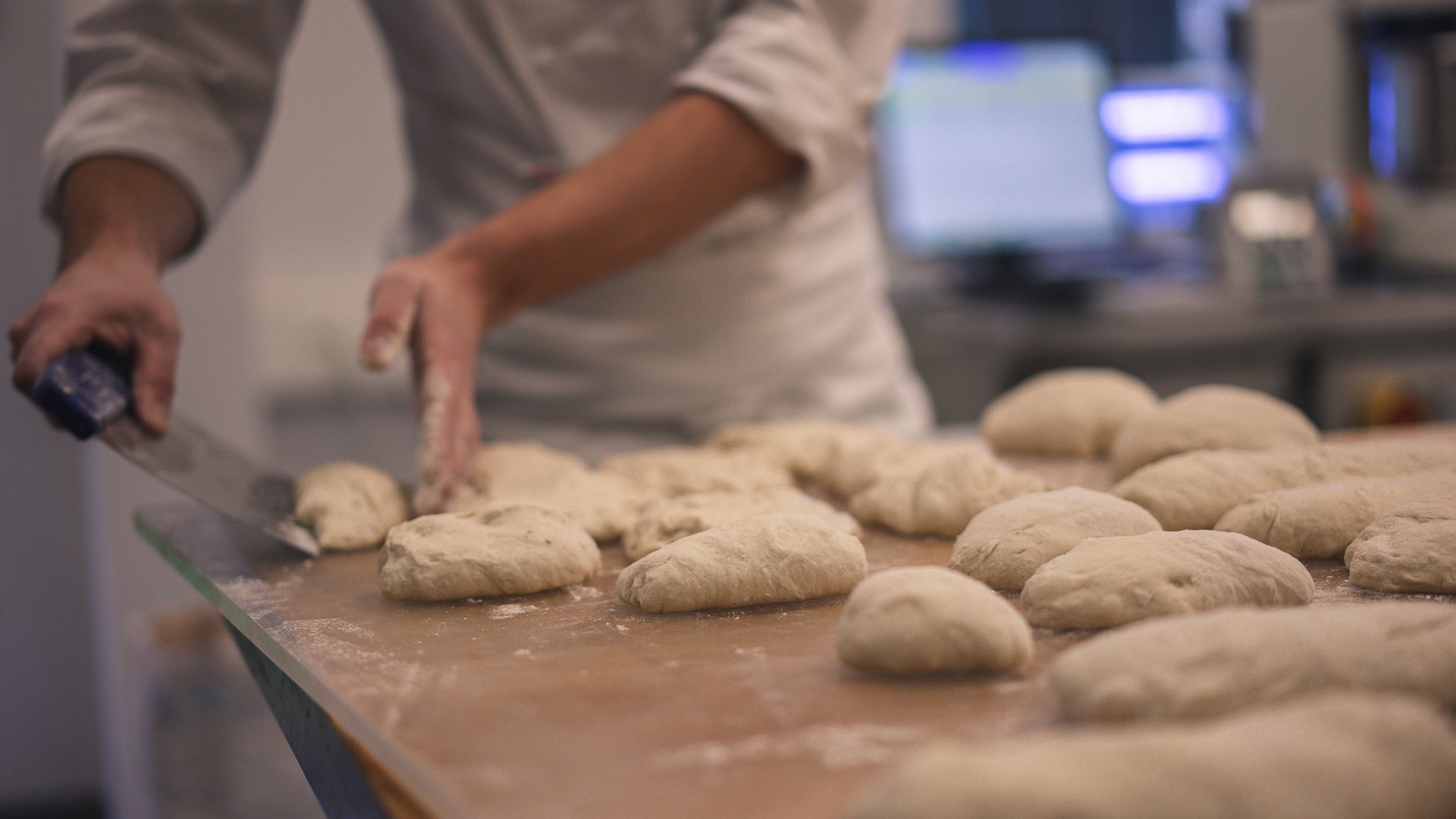 Close-up of a baker shaping and cutting fresh dough pieces on a floured surface, with a professional kitchen in the background. The focus is on the craft of bread-making.