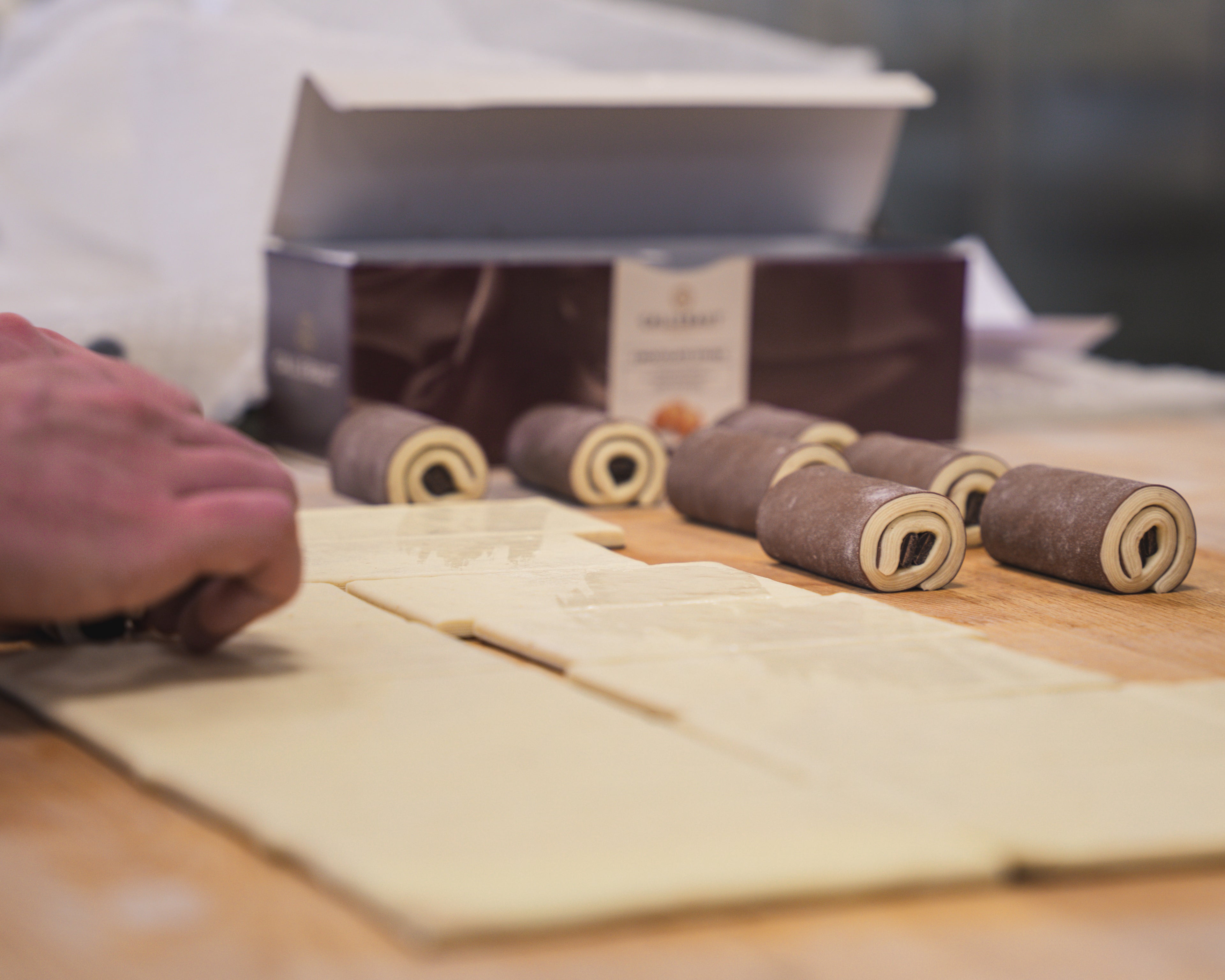 Preparation of chocolate breads on a wooden surface, with sheets of dough being cut and chocolate-filled rolls placed nearby.