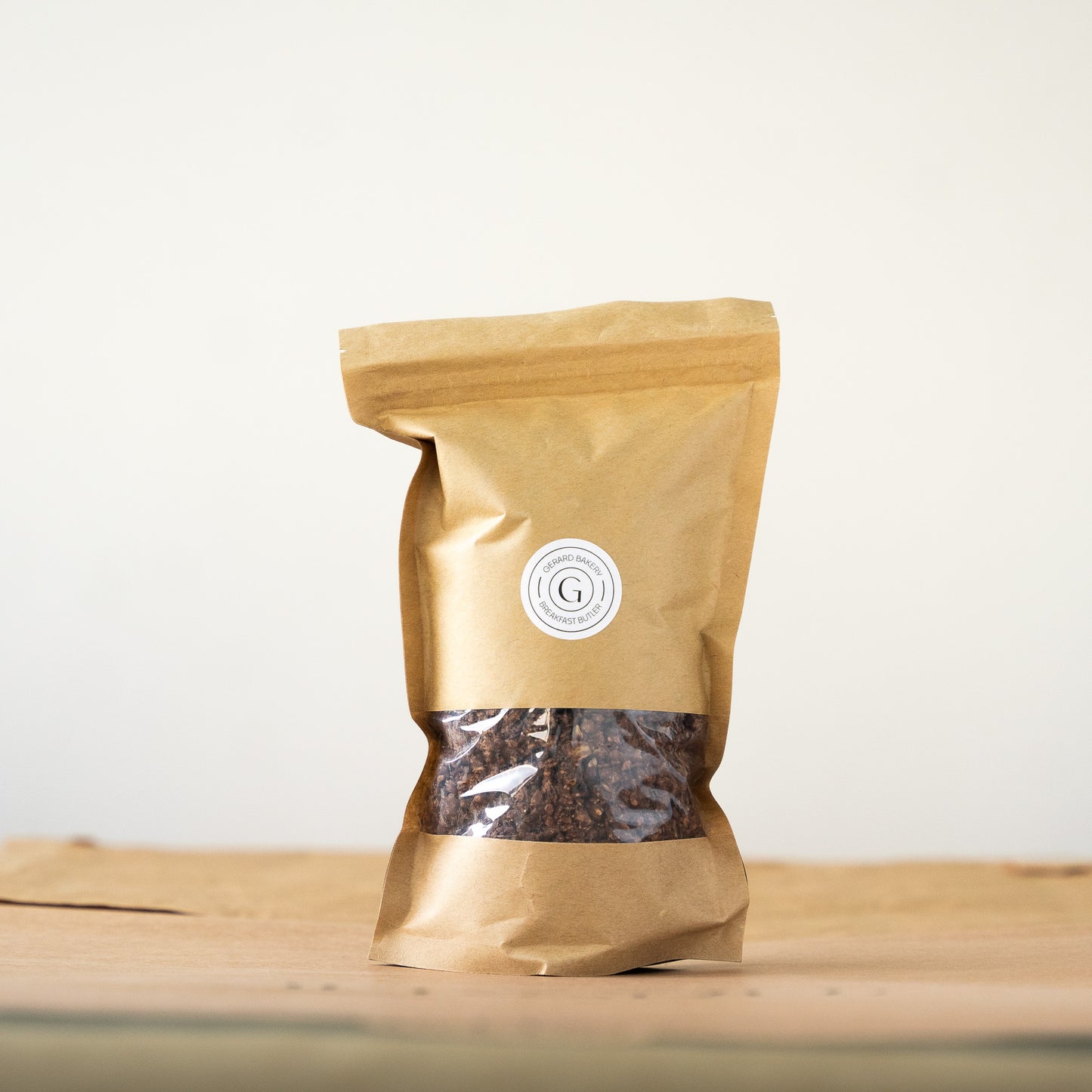 A 300-gram package of homemade chocolate granola in an elegant kraft paper bag with a transparent window, showcasing the crunchy contents, set on a light wooden surface