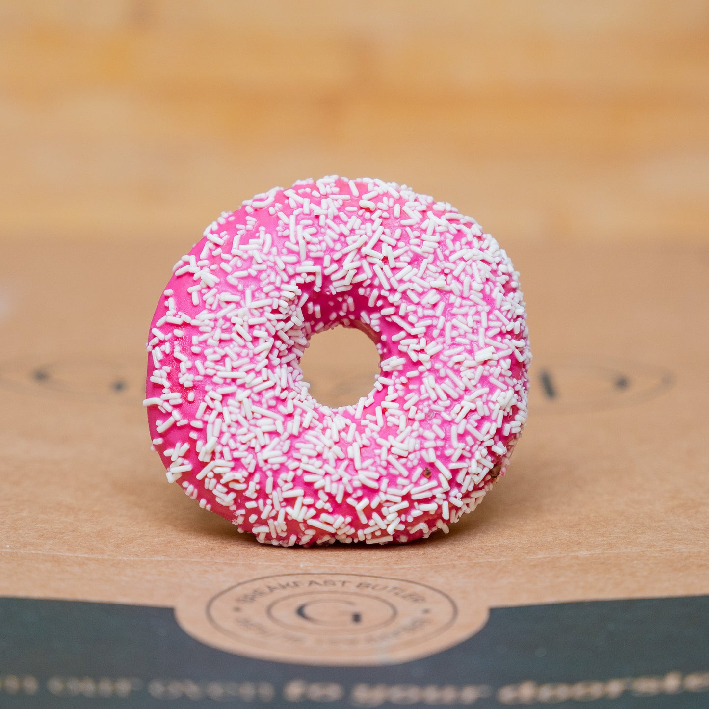 Bright pink donut topped with white sprinkles, placed on a natural brown background, showcasing its vibrant color and delicious texture