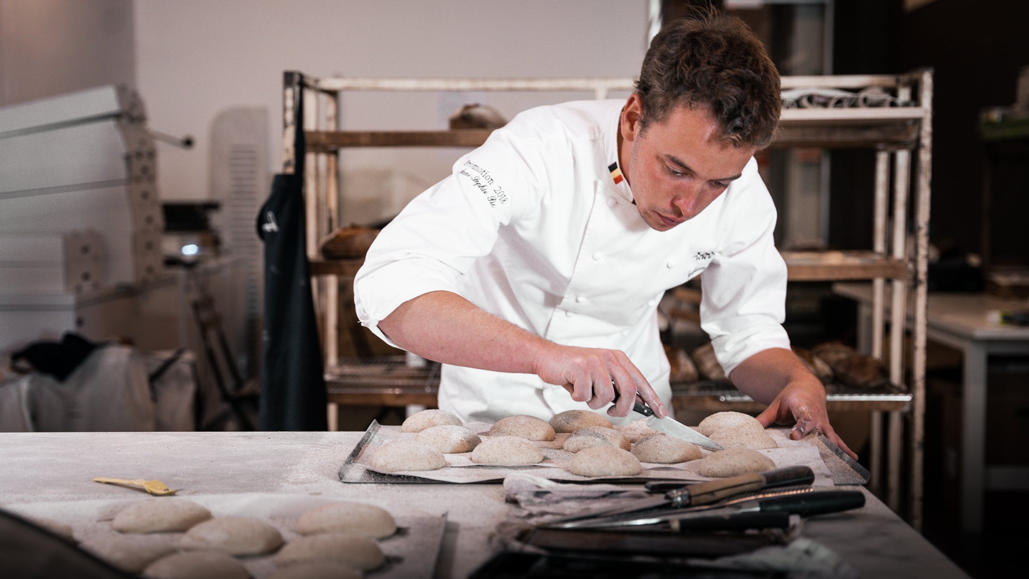 Chef Sébastion in a bakery, carefully scoring pistolets on a baking tray, with fresh dough and baking tools in the foreground.