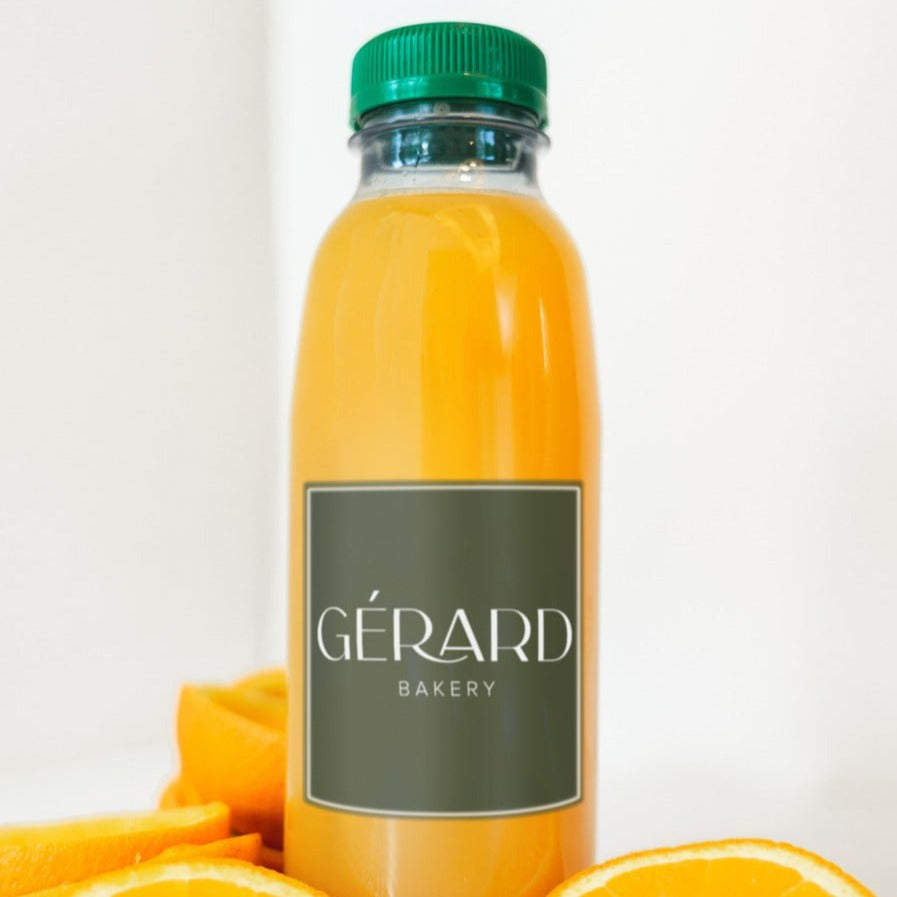 A clear bottle of freshly pressed orange juice labeled with "Gérard Bakery," featuring a green cap. The vibrant orange color of the juice is accentuated by the fresh orange slices placed at the base of the bottle, creating a refreshing and enticing visual.